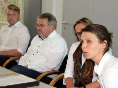 Vier Personen - zwei M&auml;nner und eine Frau - sitzen nebeneinander an Tischen, auf denen Papiere liegen. Eine der Frauen spricht.