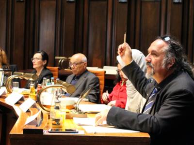 Sechs Personen, davon 4 Frauen und zwei Männer sitzen im Hodlersaal hinter Pulten. Ein Mann rechts im Bild hebt die Hand hoch, um einen Redebeitrag zu signalisieren. In der Hand hält er einen Stift.