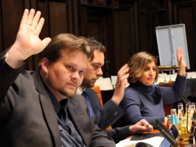 Drei Personen - eine Frau und zwei M&auml;nner - sitzen im Hodlersaal nebeneinander hinter Pulten und heben die Hand zur Abstimmung hoch.