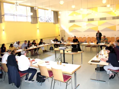 Etwa 20 Personen sitzen in einem gro&szlig;en Raum hinter rechteckig aufgestellten Tischen. Weiter hinten sitzen Zuschauer*innen auf St&uuml;hlen, die in Reihen aufgestellt sind.