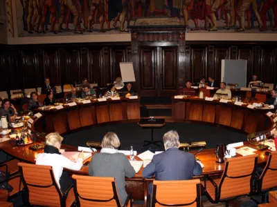 Etwa 20 Personen sitzen an kreisf&ouml;rmig aufgestellen Pulten im Hodlersaal des Neuen Rathauses.