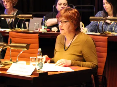 Eine Frau sitzt im Hodlersaal hinter einem Pult und spricht. In der Sitzreihe hitner ihr sind drei weitere Personen erkennbar.
