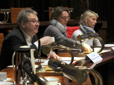 Zwei Frauen und ein Mann sitzen im Hodlersaal des Neuen Rathauses hinter Pulten. Der mann h&auml;lt eine Kaffeetasse in der Hand. Alle drei blicken nach vorn.