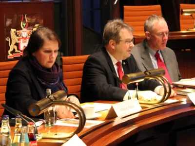 Drei Personen - eine Frau und zwei M&auml;nner - sitzen nebeneinander hinter Pulten im Hodlersaal. Die Frau schaut nach unten rechts, die M&auml;nner schauen nach vorne.