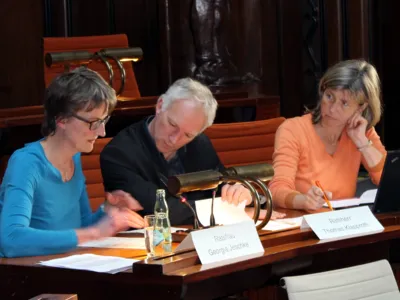 Drei Personen sitzen nebeneinander hinter Pulten. Die linke und die mittlere Personen schauen auf ein Papier, das vor der linken liegt. Die rechte schaut zur mittleren Person.