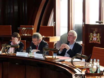 Drei Personen sitzen im Hodlersaal hinter Pulten. Die linke und die mittlere schauen auf Papiere, die vor ihnen liegen, die rechte Person schaut nach vorne.