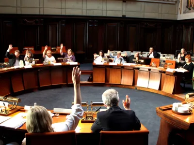 Etwa 15 Personen sitzen im Hodlersaal hinter kreisf&ouml;rmig aufgestellten Pulten. Die Personen im inneren Kreis heben die H&auml;nde zur Abstimmung.