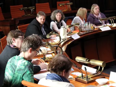 SIeben Personen sitzen im Hodlersaal des Neuen Rathauses hinter Pulten. Einige davon schauen auf Unterlagen, die vor ihnen liegen.