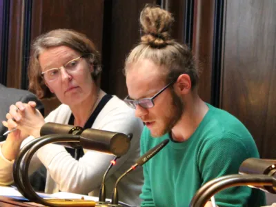 Zwei Personen sitzen im Hodlersaal hinter Pulten. Die Rechte - ein Mann - schaut auf ein Papier vor sich und spricht. Die Frau links daneben blickt zu ihm.