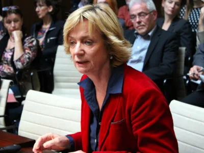 Eine Frau sitzt im Holdersaal hinter einem Pult und spricht. Hinter ihr sind etwa acht weitere Personen erkennbar.