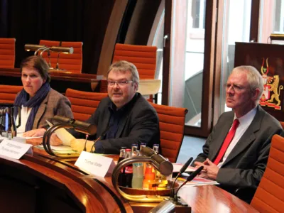 Drei Personen sitzen in Hodlersaal hinter Pulten. EIn Mann in der Mitte spricht und blickt dabei nach links.