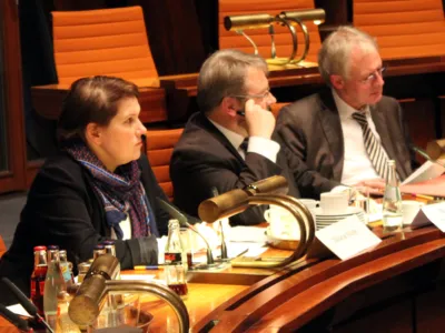 Drei Personen sitzen nebeneinander hinter Pulten. Die Frau und ein Mann blicken nach vorne, der Mann rechts blickt auf Papiere, die vor ihm auf dem Pult liegen.