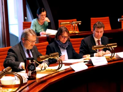 Drei Personen sitzen im Hodlersaal nebeneinander hinter Pulten und blicken auf die Unterlagen, die vor ihnen liegen.