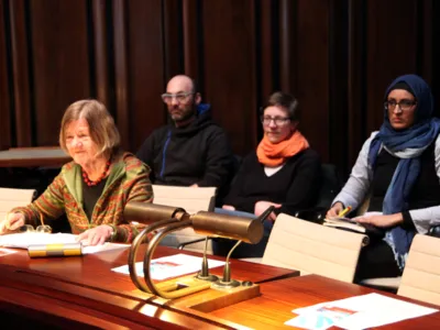 Eine Frau sitzt im Hodlersaal hinter einem Pult und spricht in ein Mikrofon. Hinter ihr sind drei weitere Personen erkennbar.