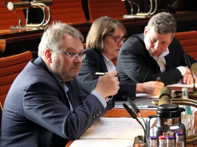 Drei Personen sitzen im Holdersaal nebeneinander hinter Pulten.