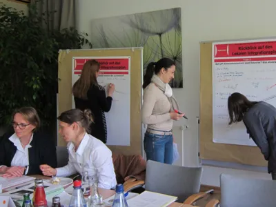 Drei Frauen stehen an zwei Plakatst&auml;ndern, auf denen sie Notizen eintragen. Davor sitzen zwei weitere Frauen an Tischen, von denen eine zur anderen spricht.