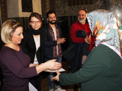 Fünf Personen stehen im Mosaiksaal des Neues Rathauses. Eine Frau überreicht einer anderen eine Urkunde und streckt ihr zur Gratulation ihre rechte Hand entgegen.