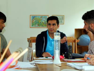EIn Jugendlicher sitzt am Kopfende eines Tisches und spricht. Links und rechts von ihm sitzen zwei weitere Jugendliche und blicken zu ihm.