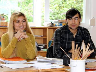 Zwei Jugendliche - eine Frau und ein Mann - sitzen nebeneinander hinter Tischen.