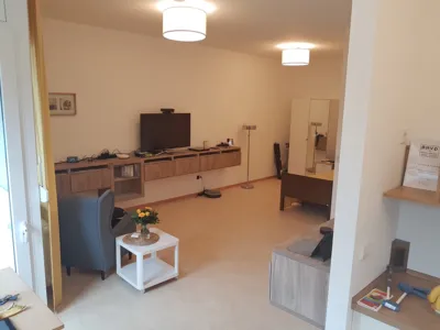 Zimmer mit Sessel, Rollcontainer und an der Wand festgeschraubten Sideboards, auf denen zahlreiche elektronische Ger&auml;te zur allt&auml;glichen Unterst&uuml;tzung liegen.