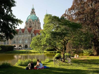 Eine Gruppe von Personen, die mit Fahrrädern unterwegs sind, picknickt auf dem Rasen am Maschteich mit Blick auf das Neue Rathaus von Hannover