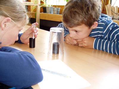 Ein Mädchen und ein Junge, die an einem Tisch sitzen, untersuchen mit Hilfe eines umgestülpten Glases klimatische Veränderungen in zwei Gefäßen