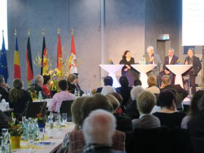 Eine Gesprächsrunde von drei Männern und einer Frau auf einem Podium, davor sitzt an langen Tischen das Publikum. Im Hintergrund sind sechs Flaggen zu sehen, unter anderem die europäische, die rumänische und die deutsche.