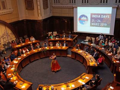 Eine indische Tänzerin mit landestypischer Kleidung steht in der Mitte des Hodlersaals, in dem Pulte ringförmig um die Mitte angeordnet sind. An den Pulten und auf den dahinter positionierten Sitzreihen sitzen indische und deutsche Teilnehmerinnen und Teilnehmer an der Veranstaltung "India Days".