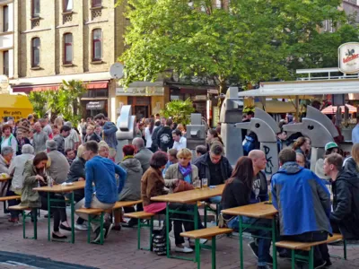 Viele Menschen stehen an einem Platz oder sitzen auf B&auml;nken an Tischen und trinken und essen.