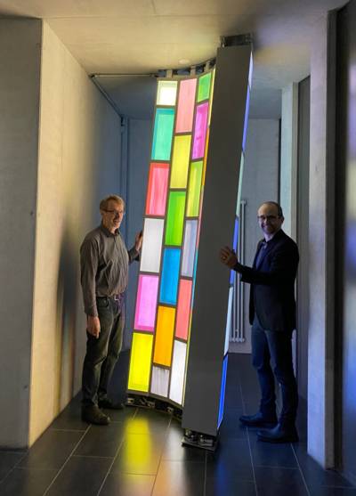 Zwei Männer an einem aus farbigen beleuchteten Elementen bestehenden hochkantigen Objekt.