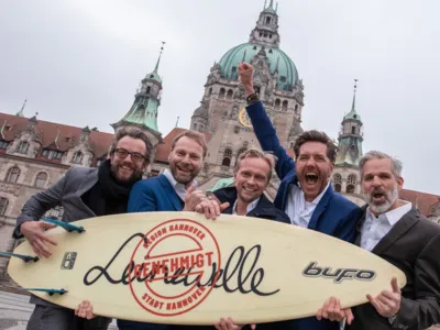 F&uuml;nf M&auml;nner halten freudestrahlend ein Surfbrett hoch, das die Aufschrift "Leinewelle genehmigt" tr&auml;gt