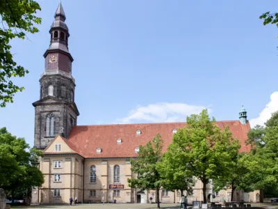 Kirche mit Kirchturm, auf dem Kirchenplatz davor stehen B&auml;ume sowie einzelne Kunstobjekte.
