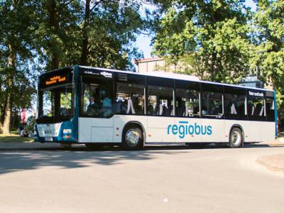 Ein Bus mit der Aufschrift regiobus stehend aus Front-/Seitenansicht, im Hintergrund eine Grünflache mit See.
