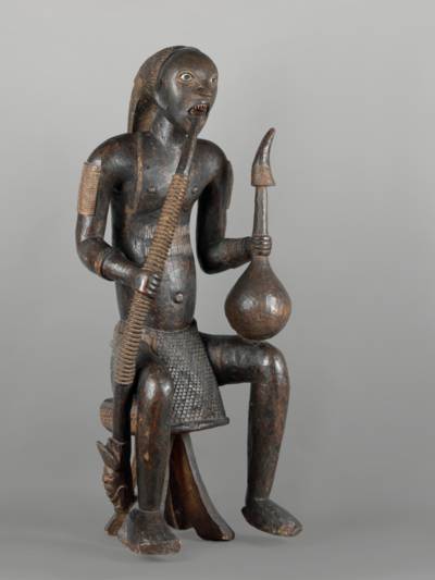 Diese Figur stellt einen König aus Bakowen im Kameruner Grasland dar, der die Symbole seines hohen Statuts trägt: geschliffene Zähne, Palmweinkalebasse mit Trinkhorn und Prunkpfeife. Er sitzt auf einem Hocker, was ebenfalls ein Zeichen seiner Würde ist.