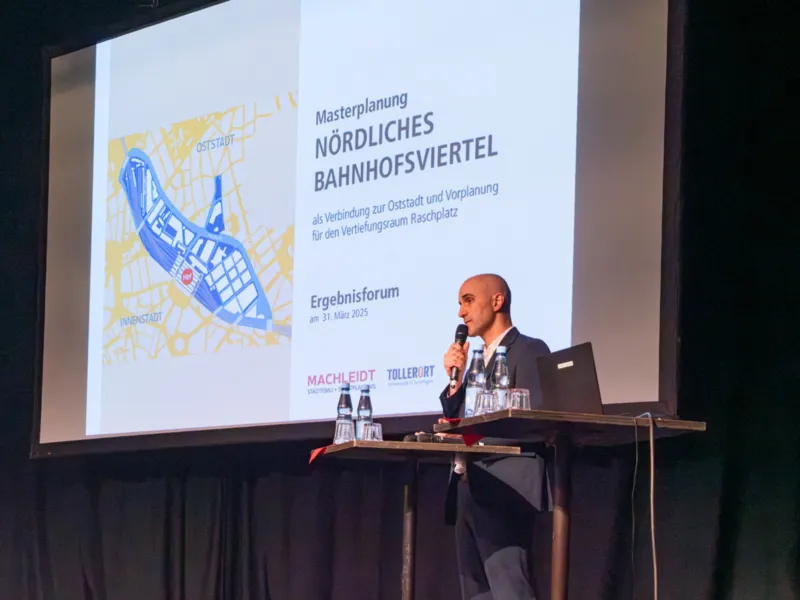Ein Mann steht mit einem Mikrofon auf einer B&uuml;hne vor einem Rednerpult, daneben eine Pr&auml;sentation auf einer Leinwand mit dem Titel "Masterplanung n&ouml;rdliches Bahnhofsviertel"
