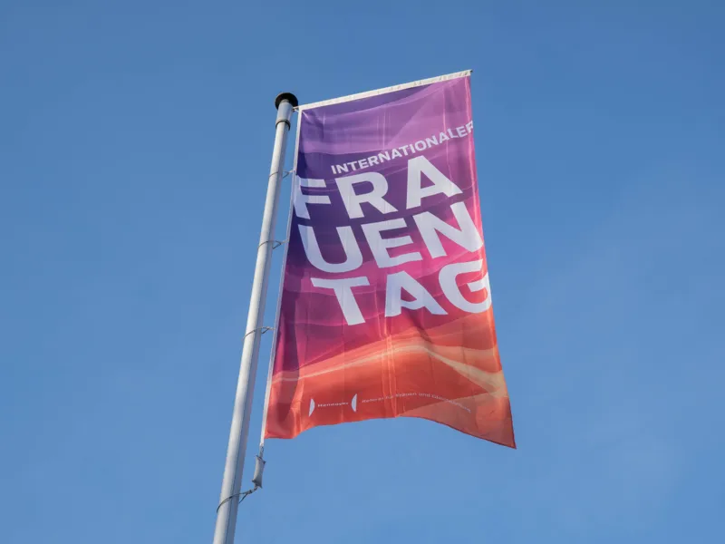 Eine Flagge im Wind vor blauem Himmel in den Farben Lila bis Pink, darauf steht Internationaler Frauentag.