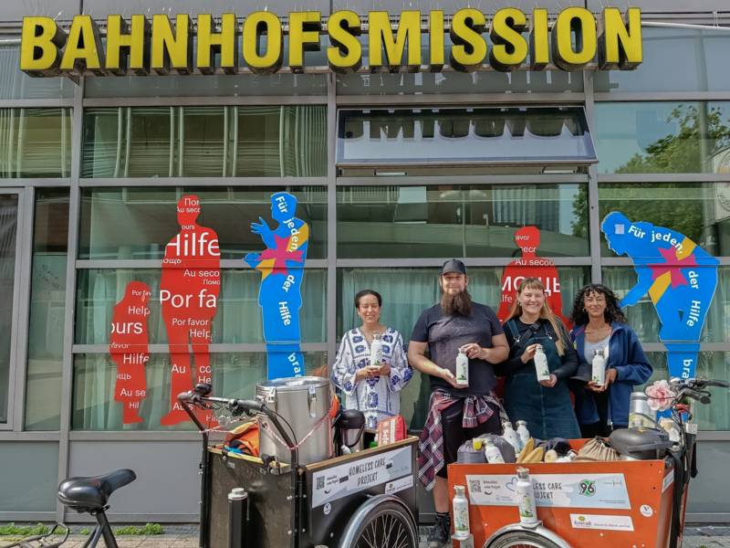 Vier Personen stehen vor einem Geb&auml;ude mit dem Text Bahnhofsmission. Sie halten Flaschen in der Hand, vor ihnen stehen zwei voll beladene Transport-Fahrr&auml;der.