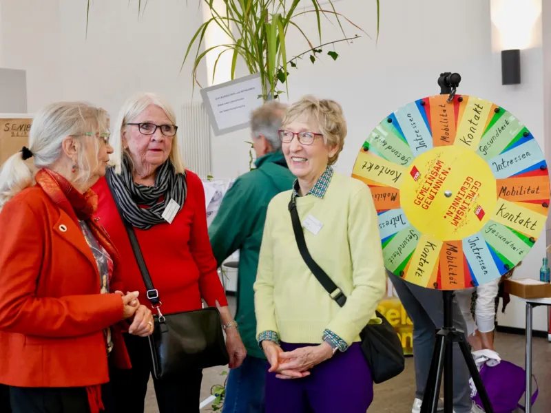 Drei Frauen unterhalten sich, im Hintergrund eine Drehscheibe mit Gewinnen wie Kontakt, Mobilit&auml;t und Interessen.