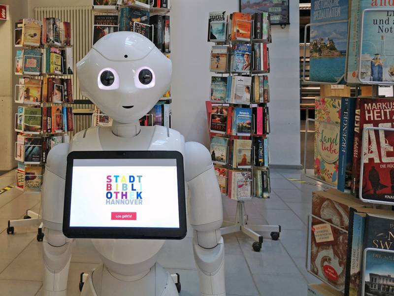 Ein Roboter steht umgeben von Bücherständern in einer Bibliothek.