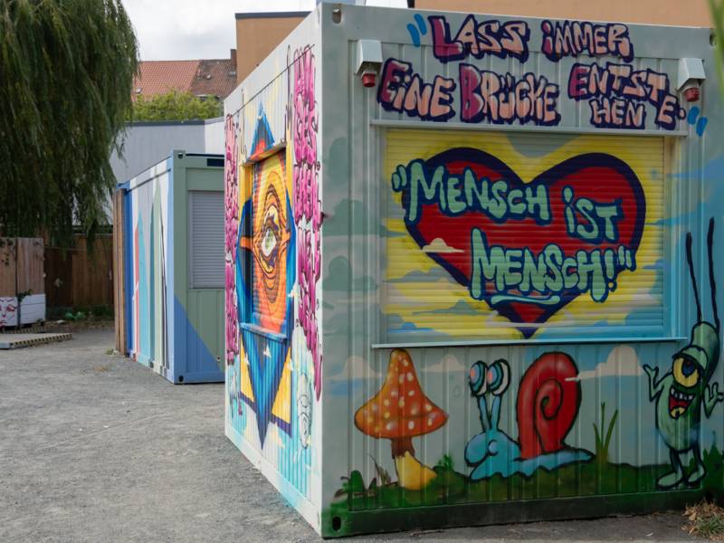 Auf einem Platz steht ein mit buntem Graffiti gestalteter Container. Darauf steht unter anderem Mensch bleibt Mensch.