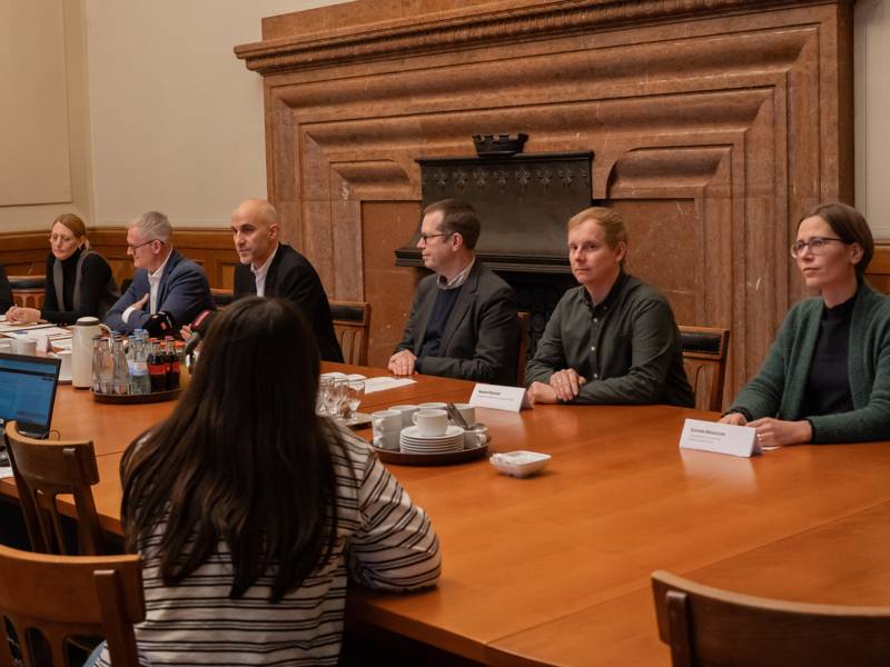 Sechs Personen sitzen an einem langen Tisch auf einer Seite, gegenüber sind Stühle zu erkennen und ein Laptop sowie eine Frau, die ihnen gegenüber sitzt.