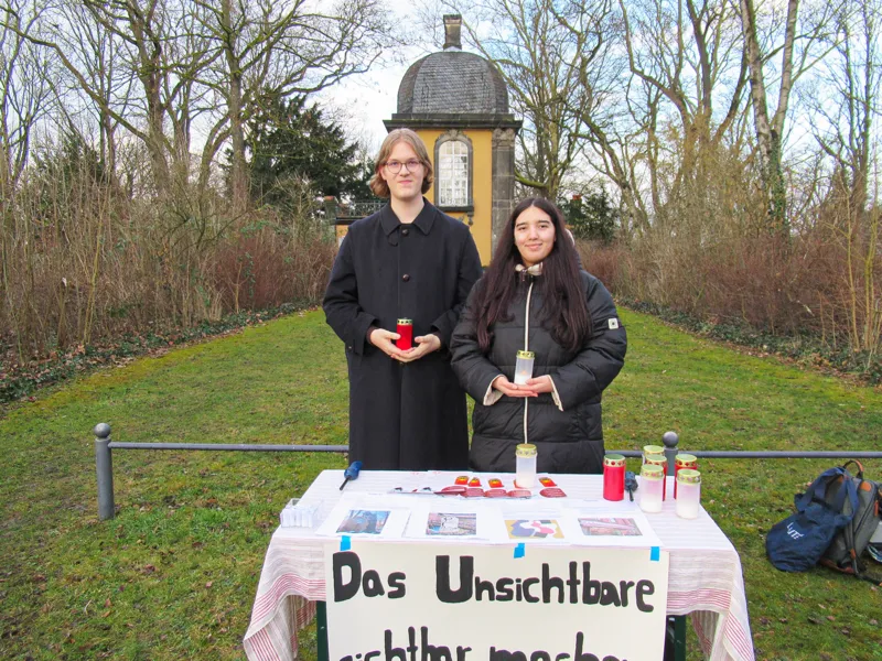 Zwei Sch&uuml;ler*innen hinter einem Tisch auf einem Stadtteilfriedhof. Darauf liegt ein Banner, auf dem steht: "Das Unsichtbare sichtbar machen".