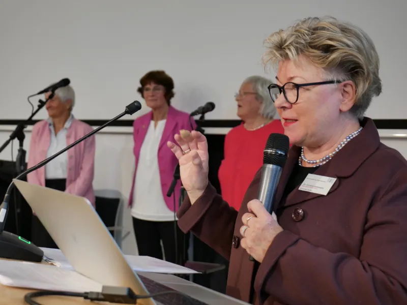 Eine Frau spricht in ein Mikrofon, seitlich stehen drei weitere Frauen vor Mikrofonen.