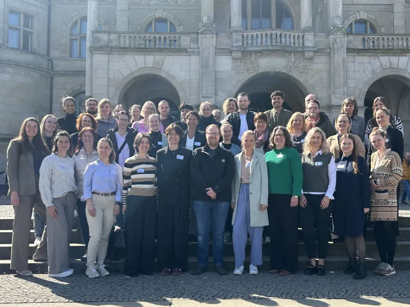 Eine gro&szlig;e Gruppe von Menschen, Frauen und M&auml;nner, stehen vor dem Neuen Rathaus und l&auml;cheln in die Kamera.