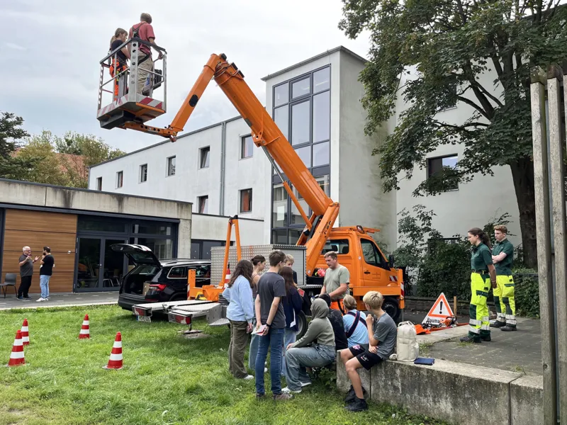 Junge Menschen bei einer Messe. Sie stehen auf der Gr&uuml;nfl&auml;che, auf der ein Hub-Fahrzeug einige in die H&ouml;he hebt.