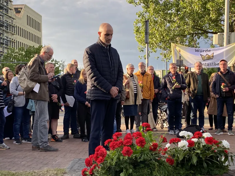 Ein Mann steht mit gesenktem Blick vor einem Blumengesteck, hinter ihm stehen Menschen.