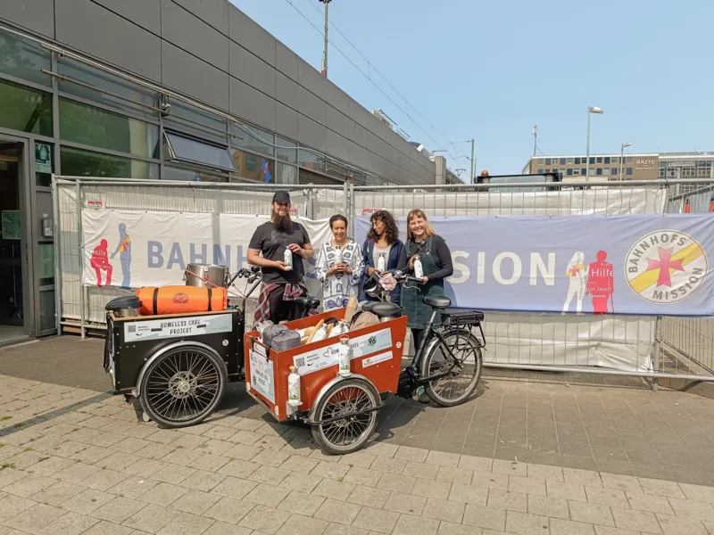 Vier Personen stehen vor einem Banner auf einem Bauzaun mit dem Text Bahnhofsmission. Sie halten Flaschen in der Hand, vor ihnen stehen zwei voll beladene Transport-Fahrr&auml;der.