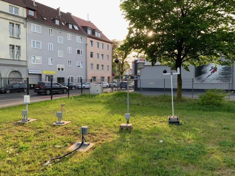 Auf einem gr&uuml;nen Rasen vor Wohnh&auml;usern und neben einer Stra&szlig;e stehen f&uuml;nf graue K&auml;sten, zwei mit hochragenden Pf&auml;hlen und Kugeln und Hauben daran.