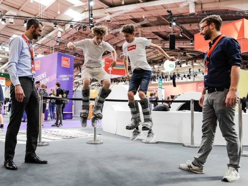 Zwei m&auml;nnliche Teenager mit stiefel&auml;hnlichen Konstruktionen an den Beinen springen in die H&ouml;he, zwei M&auml;nner schauen zu.