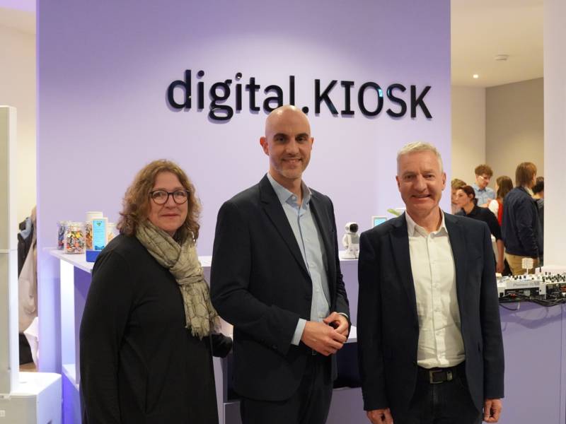 Eine Frau und zwei Männer stehen vor einer lilafarbenen Fläche mit dem Text "digital.KIOSK".
